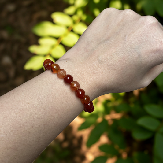 Carneool armband met warme roodbruine kralen, gedragen om de pols, gefotografeerd in natuurlijk zonlicht met zachte groene achtergrond.